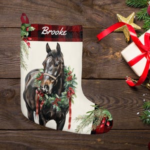 May include: Christmas stocking featuring a horse wearing a wreath. The stocking has a red and black plaid cuff with the name "Brooke" in white. The horse is brown with a wreath of greenery and red berries. A red ribbon hangs from the top.
