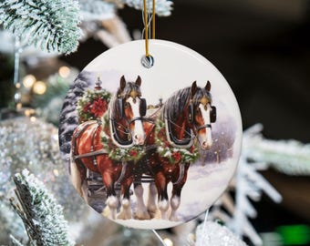 May include: Round Christmas ornament featuring a watercolor painting of two brown horses pulling a sleigh. The horses wear festive wreaths and are set against a snowy winter scene. The ornament hangs from a gold string.
