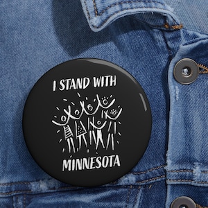 May include: A black button with white text and a graphic. The text reads "I STAND WITH MINNESOTA." The graphic depicts stylized figures. The button is set against a blue denim jacket.