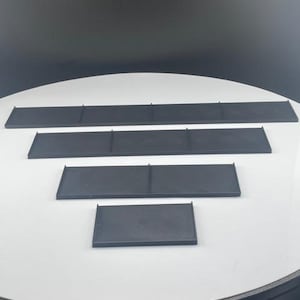 May include: Four black rectangular shelves of varying sizes are arranged on a white surface. The shelves are made of a matte material and have a simple, minimalist design. The shelves are likely for organizing or displaying items.