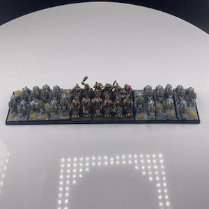 May include: A miniature tabletop game army on a black base. The figures are painted in various colors, including green, brown, and gray. The base includes rocky terrain. The figures are arranged in a line, ready for battle.