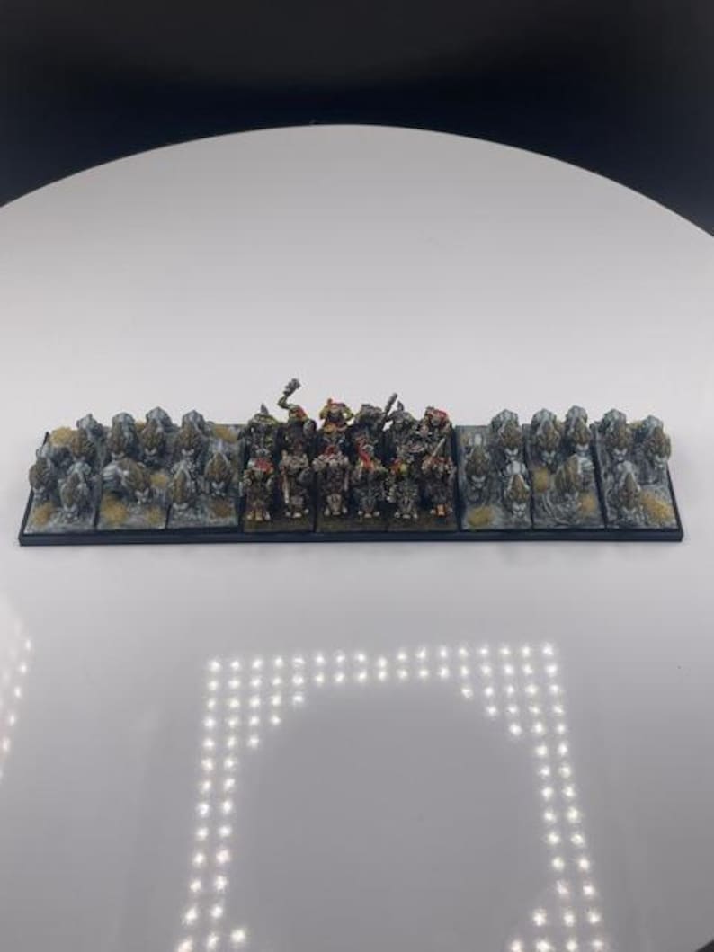 May include: A miniature tabletop game army on a black base. The figures are painted in various colors, including green, brown, and gray. The base includes rocky terrain. The figures are arranged in a line, ready for battle.
