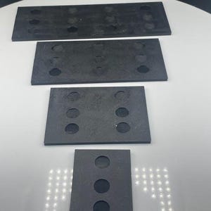May include: Four dark gray rectangular rubber pads of varying sizes are arranged vertically. Each pad has multiple circular holes. The top pad is the largest, with the bottom pad being the smallest. The pads are on a white surface.
