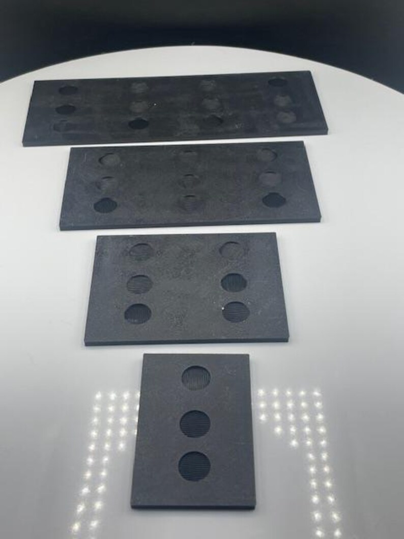 May include: Four dark gray rectangular rubber pads of varying sizes are arranged vertically. Each pad has multiple circular holes. The top pad is the largest, with the bottom pad being the smallest. The pads are on a white surface.