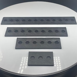 May include: Four dark gray rectangular pieces with evenly spaced circular holes. The pieces vary in size, with the largest at the top and the smallest at the bottom. The objects are arranged on a white surface.