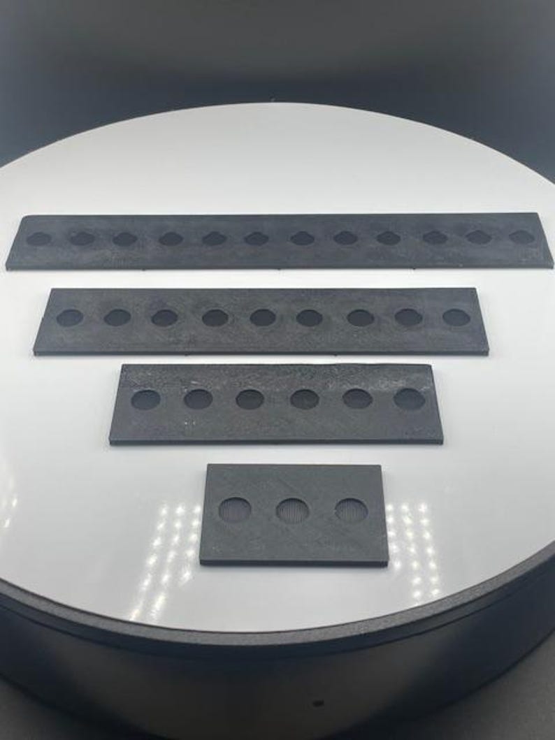 May include: Four dark gray rectangular pieces with evenly spaced circular holes. The pieces vary in size, with the largest at the top and the smallest at the bottom. The objects are arranged on a white surface.