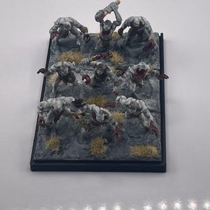 May include: Miniature tabletop game figures. The figures are of various monsters in a gray and red color scheme, posed on a rocky terrain base with patches of yellow grass. The base is black.