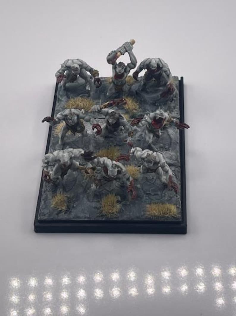 May include: Miniature tabletop game figures. The figures are of various monsters in a gray and red color scheme, posed on a rocky terrain base with patches of yellow grass. The base is black.