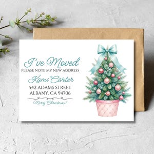 Puede incluir: Una tarjeta rectangular blanca de anuncio de mudanza con una ilustración en acuarela de un árbol de Navidad verde en una maceta rosa, rematado con un lazo azul claro y adornos rosas. El texto dice I'VE MOVED, PLEASE NOTE MY NEW ADDRESS, Kami Carter, 542 ADAMS STREET, ALBANY, CA 94706, y Merry Christmas.