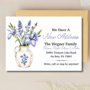 Puede incluir: Una tarjeta de anuncio de mudanza blanca con una ilustración en acuarela de flores de iris azules en una jarra de cerámica color crema. El texto dice: We Have A New Address, The Wegner Family, Shawn, Caitlin, Olivia & Miles, 16901 Treasure Lake Road, Du Bois, PA 15801, Write, call or stop by anytime!