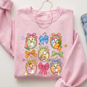May include: A light pink sweatshirt featuring a design of cartoon geese in Easter egg frames, each with a different bow. The geese are holding carrots, flowers, and eggs. A gold necklace and blue jeans are also in the image.