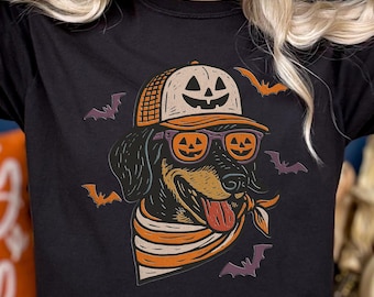 May include: Black t-shirt featuring a Halloween-themed graphic of a dog wearing sunglasses, a bandana, and a hat with a jack-o'-lantern design. The design includes bats and orange accents, perfect for the fall season.