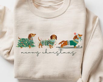 May include: Sand-colored sweatshirt featuring a festive design of three dachshunds. Each dog is adorned with holiday decorations, including a Christmas tree, lights, and a sled. The words "merry christmas" are written below the dogs.