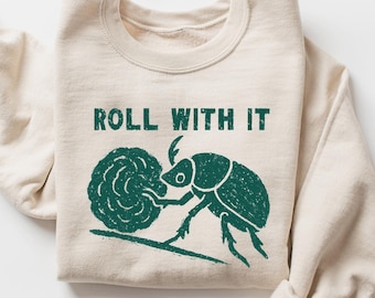 May include: Sand-colored sweatshirt with the text "ROLL WITH IT" in green. The graphic depicts a beetle rolling a ball of dung. The sweatshirt is folded with the sleeves rolled up.