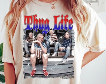 May include: A white t-shirt with the words "Thug Life" printed in red and blue letters. The shirt features a graphic of four men sitting on a porch, one of whom is wearing a red, white and blue patterned shirt.