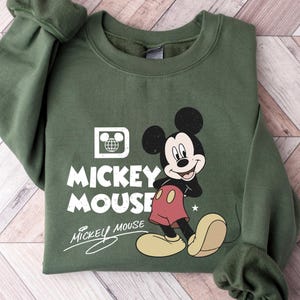 Puede incluir: Sudadera verde militar con un gr&aacute;fico de Mickey Mouse. El dise&ntilde;o incluye a Mickey, las palabras "Mickey Mouse" y un logotipo de globo. La sudadera tiene cuello redondo y mangas largas.