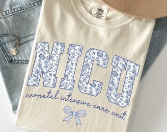 以下が含まれることがあります： 青い花柄の文字とテキストが入ったクリーム色のNICU Tシャツ。「neonatal intensive care unit」の文字が下に書かれています。Tシャツは、薄い色のジーンズ、ネックレス、ベージュの帽子の上に置かれています。