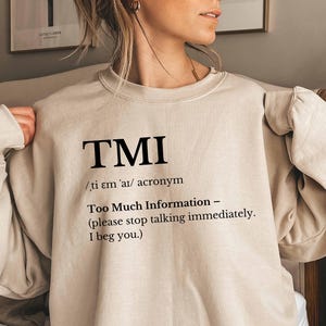 May include: Sand-colored sweatshirt with the acronym "TMI" printed in black. The text below defines "TMI" as "Too Much Information" and includes a request to stop talking. The sweatshirt is a neutral color and has long sleeves.
