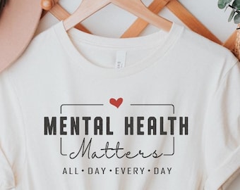 May include: A white t-shirt with the text "Mental Health Matters" in black lettering with a red heart above the text. The text "All Day Every Day" is below the main text in smaller black lettering.