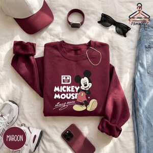 Puede incluir: Sudadera granate con un gr&aacute;fico de Mickey Mouse y el texto "Mickey Mouse". La imagen tambi&eacute;n incluye una gorra de b&eacute;isbol granate, gafas de sol, un reloj, un collar dorado, un tel&eacute;fono granate y zapatillas deportivas.