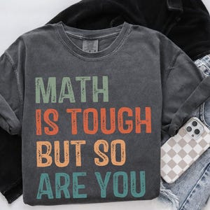 May include: A dark gray t-shirt with a vintage-style graphic that reads "MATH IS TOUGH BUT SO ARE YOU". The text is in a rainbow of colors, including red, orange, yellow, green, blue, and purple.
