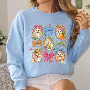 May include: Light blue sweatshirt featuring a whimsical Easter-themed design. The design includes cartoon ducks in ornate frames, surrounded by colorful bows, carrots, and Easter eggs. Butterflies and floral accents add to the festive look.