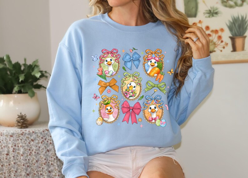 May include: Light blue sweatshirt featuring a whimsical Easter-themed design. The design includes cartoon ducks in ornate frames, surrounded by colorful bows, carrots, and Easter eggs. Butterflies and floral accents add to the festive look.