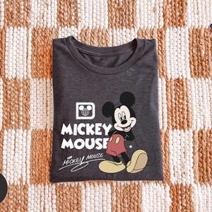 Puede incluir: Camiseta gris oscuro con un dise&ntilde;o de Mickey Mouse. El dise&ntilde;o incluye las palabras "Mickey Mouse" en blanco, con Mickey en su pose cl&aacute;sica. La camiseta est&aacute; doblada sobre una superficie estampada.