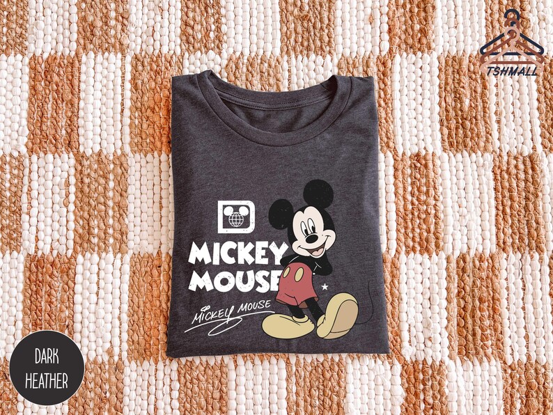 Puede incluir: Camiseta gris oscuro con un dise&ntilde;o de Mickey Mouse. El dise&ntilde;o incluye las palabras "Mickey Mouse" en blanco, con Mickey en su pose cl&aacute;sica. La camiseta est&aacute; doblada sobre una superficie estampada.