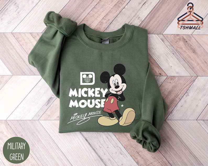 Puede incluir: Sudadera verde militar con un gr&aacute;fico de Mickey Mouse. El dise&ntilde;o incluye a Mickey, las palabras "Mickey Mouse" y un logotipo de globo. La sudadera tiene cuello redondo y mangas largas.