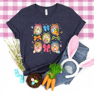 May include: A navy blue t-shirt featuring a colorful Easter-themed design with cartoon chicks in decorated eggs, bows, and butterflies. The image includes a pair of denim jeans, bunny ears, carrots, and a nest with eggs.