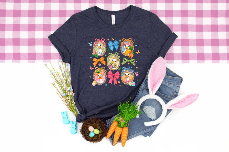 May include: A navy blue t-shirt featuring a colorful Easter-themed design with cartoon chicks in decorated eggs, bows, and butterflies. The image includes a pair of denim jeans, bunny ears, carrots, and a nest with eggs.