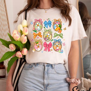 May include: Cream-colored t-shirt featuring a grid of cartoon duck illustrations with Easter-themed elements. Each duck is framed with a decorative border and adorned with bows, flowers, and Easter eggs. The shirt is paired with light blue jeans.