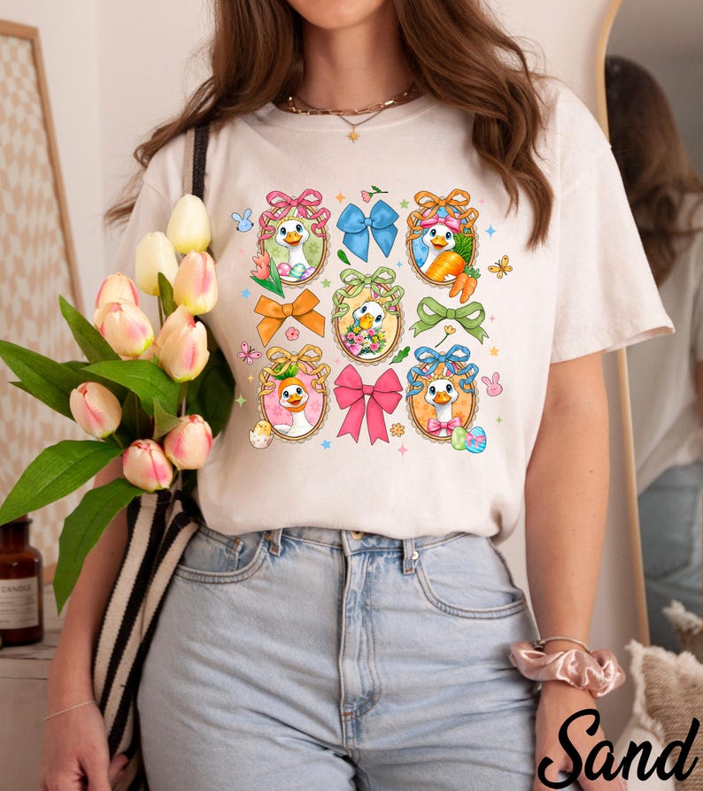 May include: Cream-colored t-shirt featuring a grid of cartoon duck illustrations with Easter-themed elements. Each duck is framed with a decorative border and adorned with bows, flowers, and Easter eggs. The shirt is paired with light blue jeans.