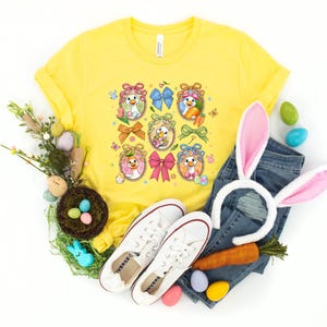 May include: A yellow t-shirt with a colorful Easter-themed graphic featuring cartoon ducks in egg-shaped frames, bows, and carrots. The image includes a pair of white sneakers, blue jeans, and a bunny ear headband.