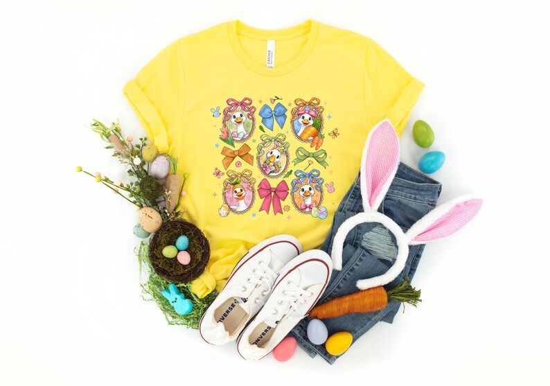 May include: A yellow t-shirt with a colorful Easter-themed graphic featuring cartoon ducks in egg-shaped frames, bows, and carrots. The image includes a pair of white sneakers, blue jeans, and a bunny ear headband.