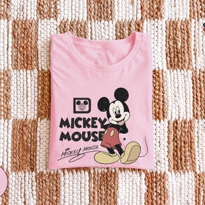 Puede incluir: Una camiseta rosa con un dise&ntilde;o de Mickey Mouse. El dise&ntilde;o incluye la imagen de Mickey Mouse, las palabras "Mickey Mouse" y una firma. La camiseta est&aacute; doblada y colocada sobre una superficie estampada.