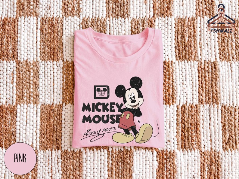 Puede incluir: Una camiseta rosa con un dise&ntilde;o de Mickey Mouse. El dise&ntilde;o incluye la imagen de Mickey Mouse, las palabras "Mickey Mouse" y una firma. La camiseta est&aacute; doblada y colocada sobre una superficie estampada.