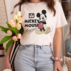 Puede incluir: Camiseta color crema con un dise&ntilde;o cl&aacute;sico de Mickey Mouse. El dise&ntilde;o incluye a Mickey Mouse en pose de caminar, las palabras "Mickey Mouse" en negrita y una firma. La camiseta se combina con vaqueros de lavado claro.