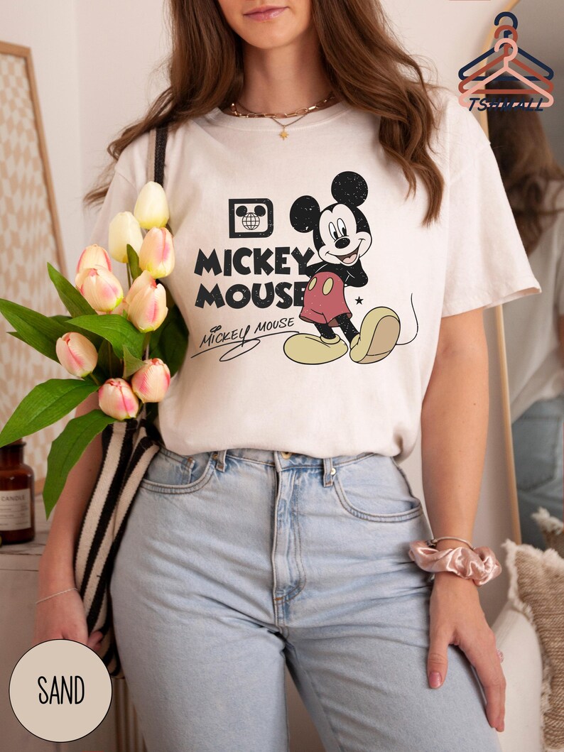 Puede incluir: Camiseta color crema con un dise&ntilde;o cl&aacute;sico de Mickey Mouse. El dise&ntilde;o incluye a Mickey Mouse en pose de caminar, las palabras "Mickey Mouse" en negrita y una firma. La camiseta se combina con vaqueros de lavado claro.