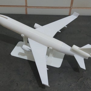 Puede incluir: Un modelo de avión de plástico blanco con un soporte. El avión tiene un fuselaje largo y estrecho y dos alas. Las alas están ligeramente inclinadas hacia arriba. El avión está sentado en un soporte de plástico blanco.