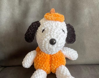 Op de afbeelding: Een handgemaakt pluche hondenspeelgoed met een witte body, zwarte oren en een zwarte knoopneus. De hond draagt een oranje outfit en een bijpassende hoed met een kleine steel, die op een pompoen lijkt. Het speelgoed staat op een grijze ondergrond.