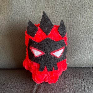 Op de afbeelding: Een knuffel gemaakt van felrood garen met een getextureerd, pluizig oppervlak. Het heeft een zwart viltmasker met scherpe, puntige bovenranden en een schedelachtige vorm. Het masker bevat twee amandelvormige ogen met witte centra en rode omlijningen.