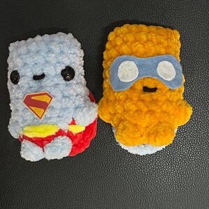 May include: Two handmade plush toys. One is light blue with a red and yellow emblem and red accents. The other is yellow with blue eye coverings. Both have black button eyes and are made of a soft, textured material.