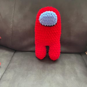May include: A handmade red crochet Among Us character with a light blue visor. The plush toy has a rounded rectangular shape with two small feet. The character is made of soft yarn.