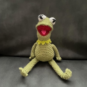 May include: A handmade crochet frog doll in shades of green, with white and black eyes, a red mouth, and a pink tongue. The frog has a yellow collar and is sitting with its legs spread. The doll is made of yarn.