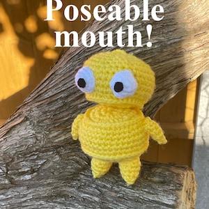 Könnte beinhalten: Eine handgefertigte gelbe Häkelfigur mit großen weißen und schwarzen Augen, kleinen Armen und Beinen. Der Text "Poseable mouth!" ist in Weiß dargestellt. Die Figur wird auf einem Holzstück präsentiert.