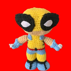 May include: A crocheted plush toy of a superhero character. The toy is yellow with black eyes and a blue and yellow costume. The toy is standing on a red background.