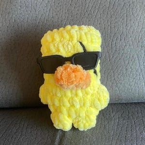 May include: A small, yellow, crocheted plush duckling made from soft, textured yarn. The duckling features a bright orange, rounded beak and is wearing a pair of black, rectangular-framed sunglasses that cover its eyes.