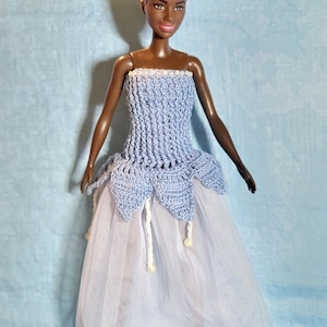 Könnte beinhalten: Eine Puppe trägt ein hellblaues Häkelkleid mit einem Tüllrock. Das Kleid hat einen weißen Häkelbesatz und weiße Perlen.